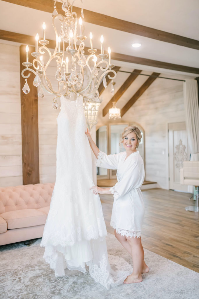 Bride smiles while posing with her high neck lace gown that is hung from the chandelier in the bridal suite