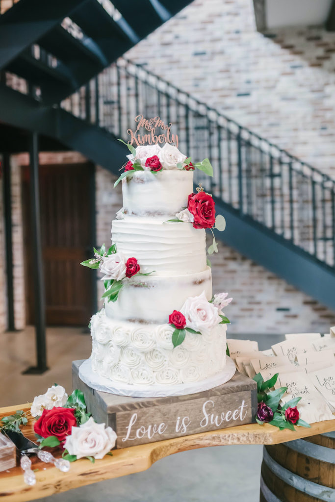 Four tier white wedding cake adorned with red and pink roses in the reception area of the Weinberg at Wixon Valley