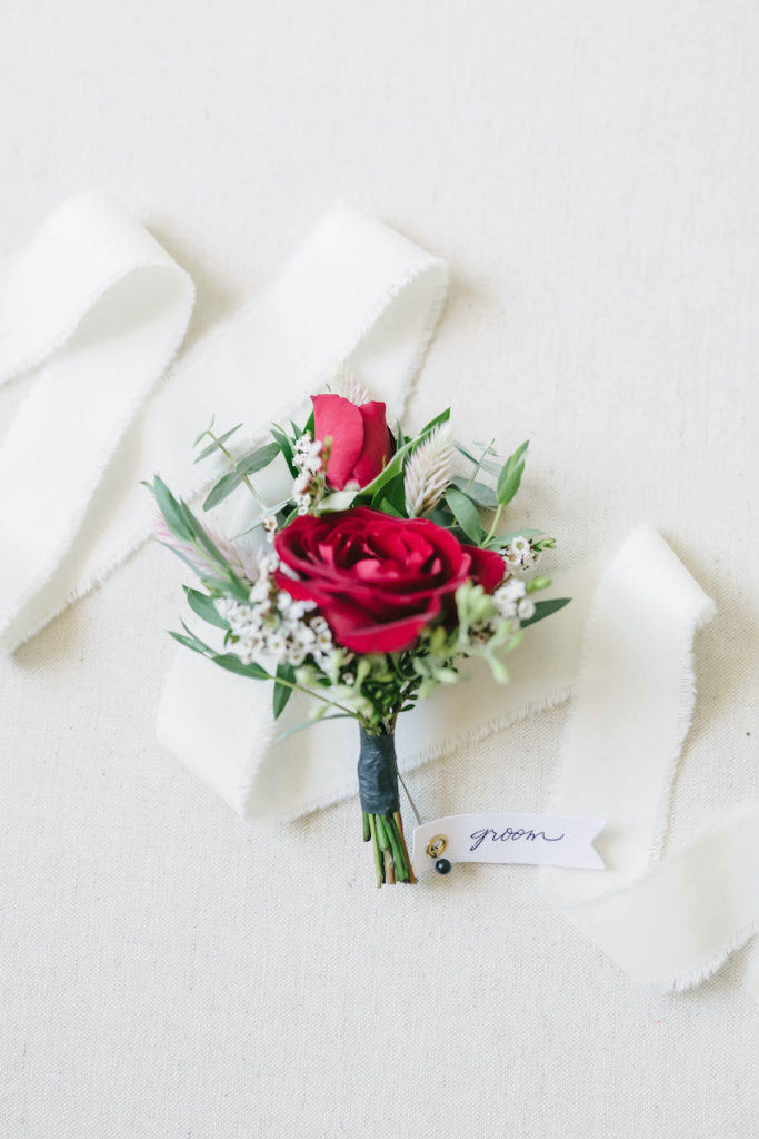 Red floral boutonniere with groom pinned laying on ivory silk ribbon