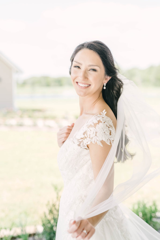 Bride with hair pulled half back and long veil hanging down from hair with a short sleeve sheer wedding dress in Alvin, TX by Christina Elliot Photography. hair inspo for bridals how should i do my hair for bridals simple wedding hair easy bridal hair half dos for bridals hair styles for veils short sleeve wedding dresses lace and sheer wedding dress #houstontexasphotographer #alvintxweddingphotographer #texasweddingphotography #outdoorbridalformals #bridalphotography
