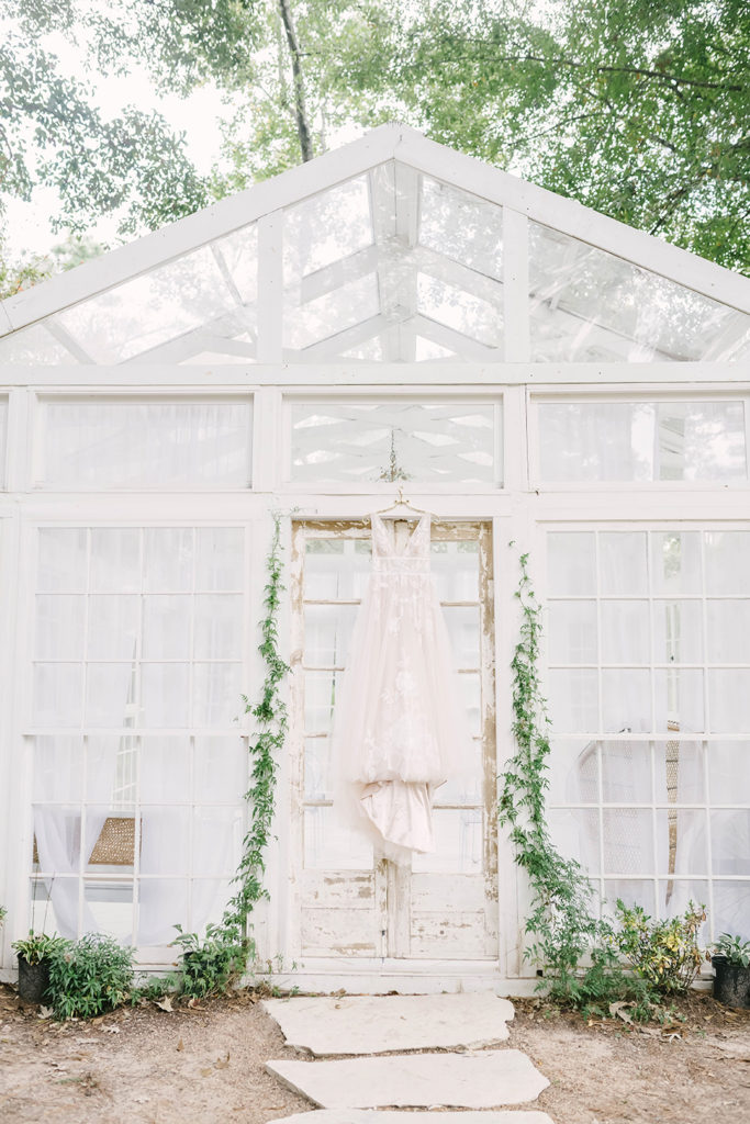 Gorgeous lace wedding dress in cream hanging on greenhouse door with leaf vines hanging on door in this wedding in Houston, TX by Christina Elliot Photography. lace wedding dress cream wedding dress wedding dress inspo sleeveless wedding dresses summer weddings dresses greenhouse wedding styling wedding dress for wedding shoot green vines leaves wedding decor #houstontexasphotographer #houstonweddingphotographer #texasweddingphotography #greenhousewedding #outdoorweddinginspo