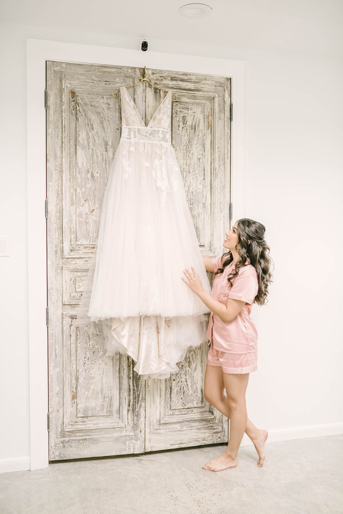 Happy bride looking at beautiful cream and lace wedding dress wearing pink bridal pajamas before she puts dress on in Houston, TX by Christina Elliot Photography. bridal outfits to get ready in cute outfits for before the wedding matching outfits for bride and bridesmaids lace cream wedding dress inspo wedding dress ideas vneck wedding dresses #houstontexasphotographer #houstonweddingphotographer #texasweddingphotography #greenhousewedding #outdoorweddinginspo