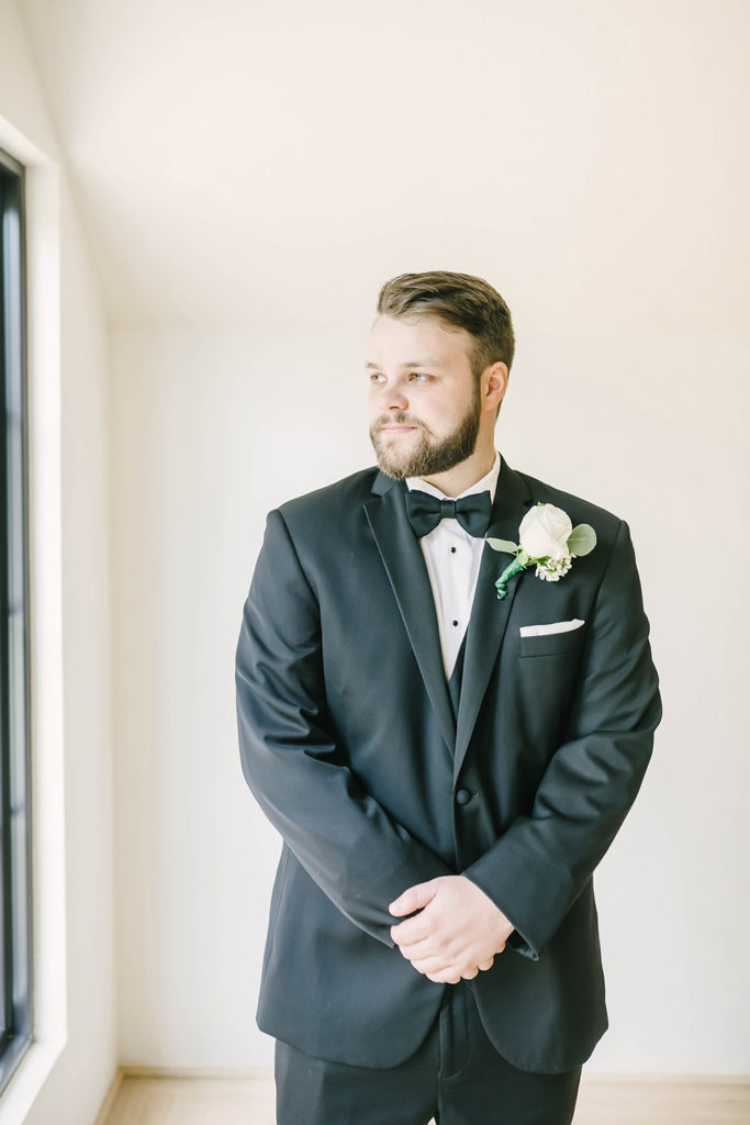 Classic groom in black tux with a black bowtie wearing a single white rose for a boutonniere by Christina Elliot Photography in Houston, TX. classic grooms how to dress your groom tux for groom tux vs suit for groom black tux for groom bowtie for groom tie vs bowtie groom boutonniere inspo houston texas wedding photographer greenhouse wedding #houstontexasphotographer #houstonweddingphotographer #texasweddingphotography #greenhousewedding #outdoorweddinginspo