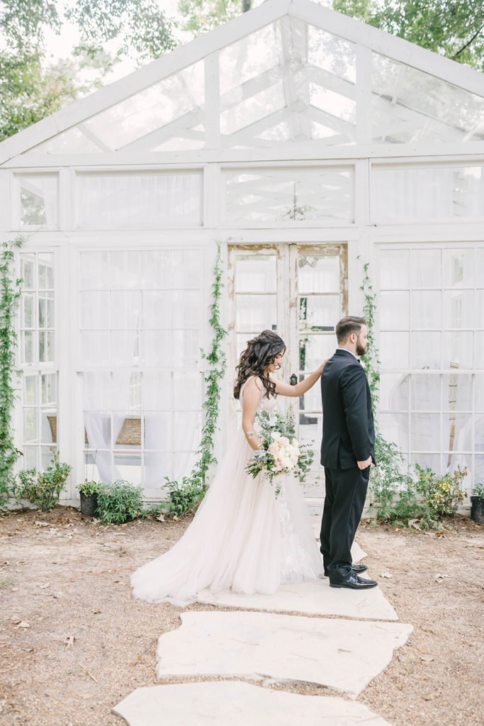 Bride and groom formals at greenhouse location with green vines floral doing first look moment in Houston, TX by Christina Elliot Photography. first look photography bride and groom first look greenhouse wedding vines and greenery decor for wedding the oak atelier woodlands houston wedding photographer cream wedding dress black tuxedo for groom #houstontexasphotographer #houstonweddingphotographer #texasweddingphotography #greenhousewedding #outdoorweddinginspo