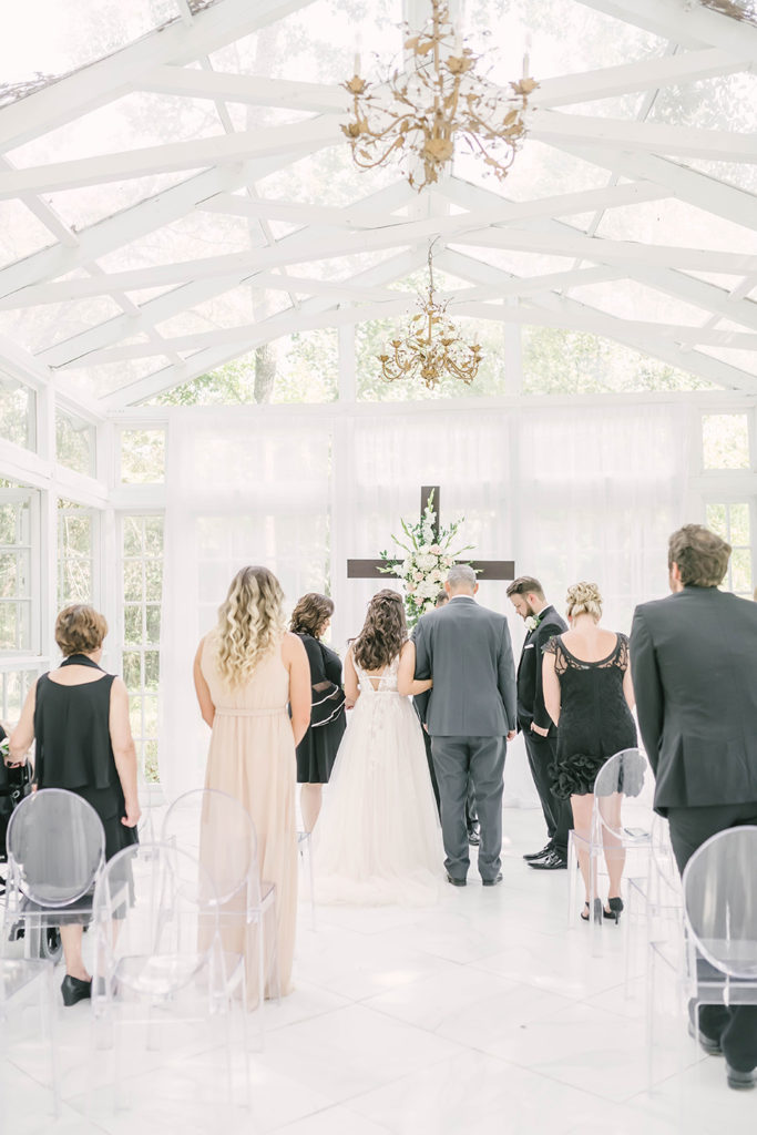 Groom and bride walking down the wedding aisle in a beautiful greenhouse with white and gold chandelier and large cross by Christina Elliot Photography in Houston, TX. wedding aisle walking down the aisle gold chandelier large cross for wedding colors white wedding the oak atelier tx greenhouse houston wedding photography woodlands #houstontexasphotographer #houstonweddingphotographer #texasweddingphotography #greenhousewedding #outdoorweddinginspo