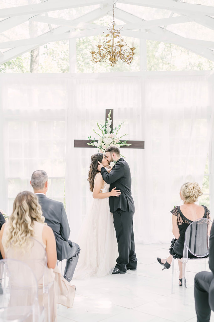 Bride and groom at altar with a large wood cross decorated with white flowers in Houston, TX by Christina Elliot Photography. wedding altar inspo how to decorate wedding altar cross at wedding catholic wedding decor inspo wedding ceremony inpso how to decorate for wedding ceremony houston wedding photography greenhouse wedding ideas #houstontexasphotographer #houstonweddingphotographer #texasweddingphotography #greenhousewedding #outdoorweddinginspo