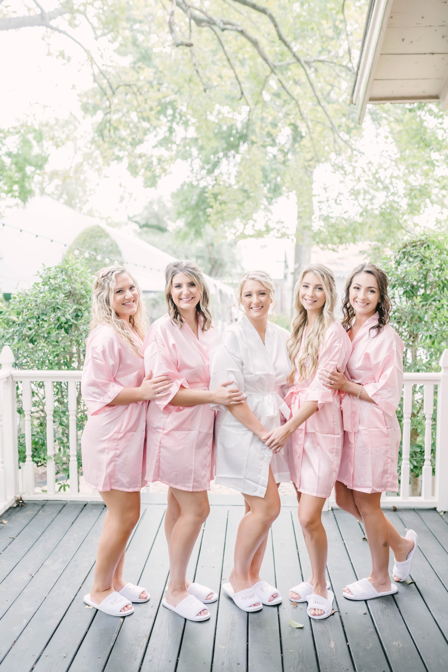 Bride poses with bridesmaids in pink and white silk robes outside of the bridal cottage