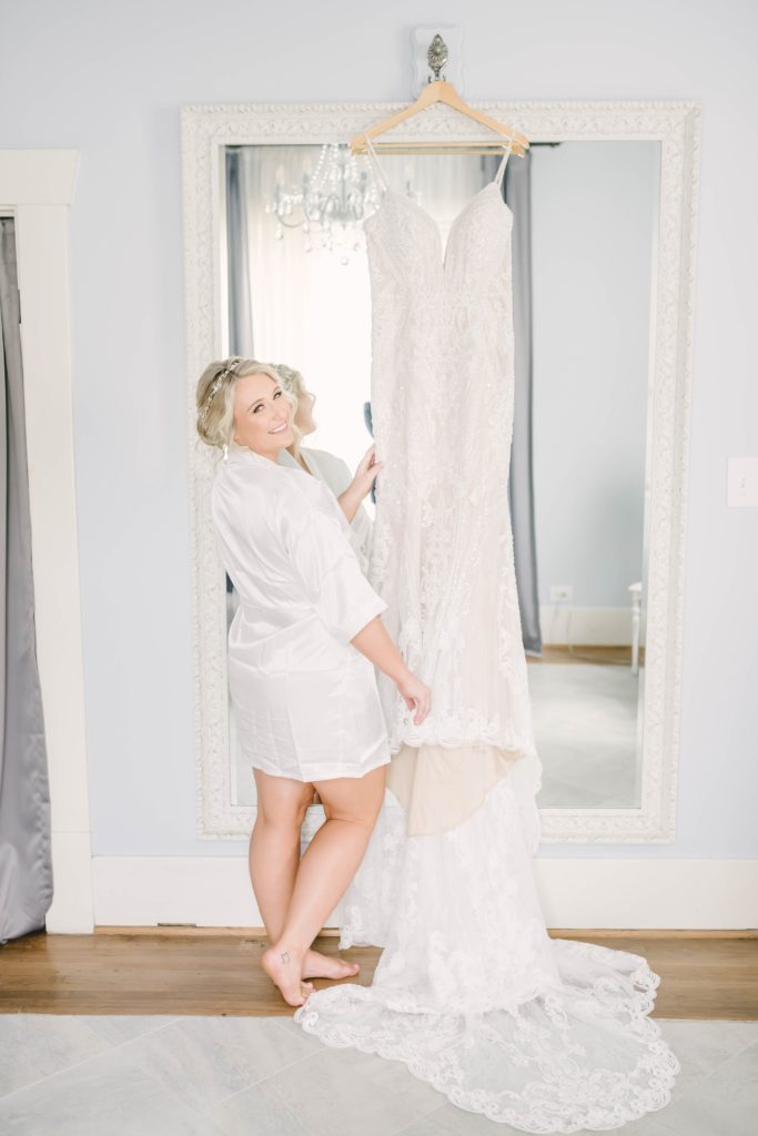 Darling photo of the bride standing next to her hanging wedding dress as she smiles for the camera before getting dressed taken by Christina Elliott. Getting ready photos wedding dress inspo shots of the bride summer weddings sleeveless wedding dress inspo