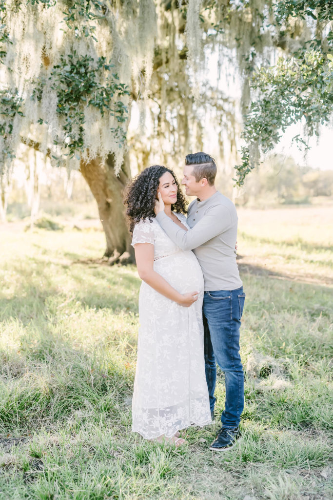 While posed under a shade tree, Christina Elliott Photography captures this young husband cupping his wife's chin romantically. tender couples poses Houston Texas maternity session photographer white and gray family photo outfit colors #HoustonMaternityPhotographer #HoustonCouplesPhotographer #HoustonPhotographer #MaternitySession #MaternityDresses #ChristinaElliottPhotography #HoustonFamilyPhotographer #Pregnant #BabyBump #WereExpecting