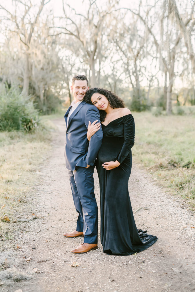 During this formal maternity session along an unpaved pathway in Houston, Texas, Christina Elliott Photography captures this couple leaning in together. pregnant woman leaning head on husband's shoulder formal maternity session Houston photographer black velvet off the shoulder maternity dress #HoustonMaternityPhotographer #HoustonCouplesPhotographer #HoustonPhotographer #MaternitySession #MaternityDresses #ChristinaElliottPhotography #HoustonFamilyPhotographer #Pregnant #BabyBump #WereExpecting