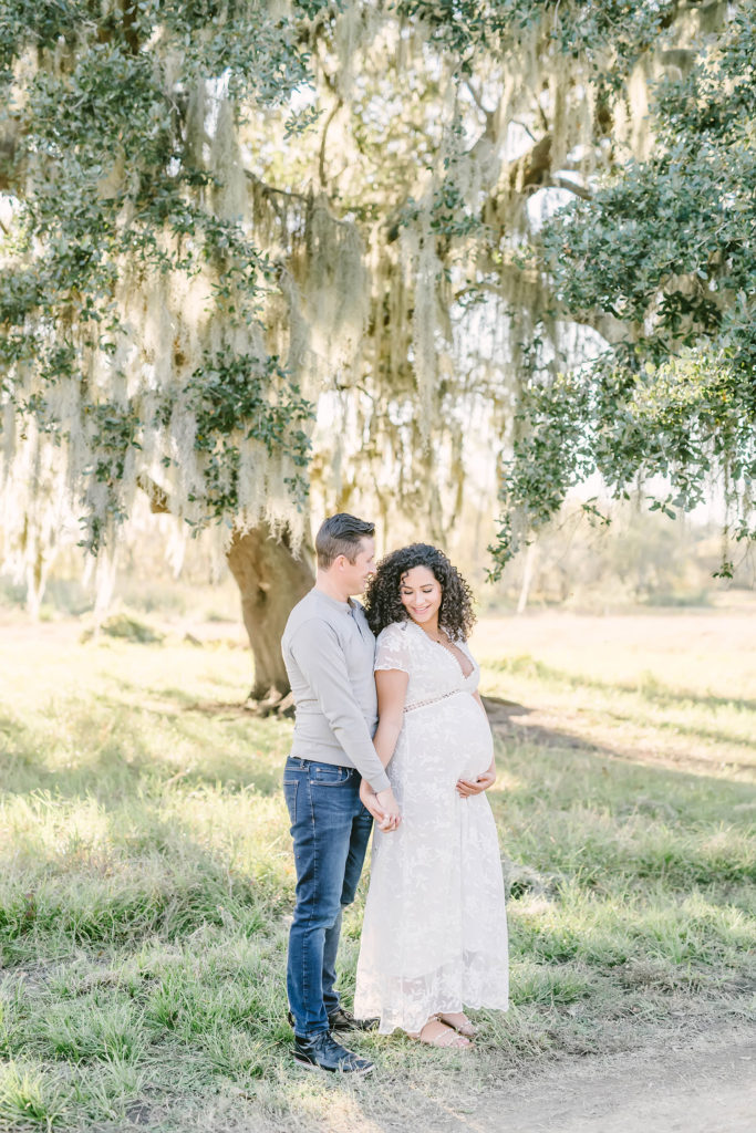 While posed under a shade tree, Christina Elliott Photography captures this expectant couple hugging from behind during this maternity session in Houston, Texas. winter Houston Texas maternity session photographer pregnant woman cupping baby bump white maternity dress #HoustonMaternityPhotographer #HoustonCouplesPhotographer #HoustonPhotographer #MaternitySession #MaternityDresses #ChristinaElliottPhotography #HoustonFamilyPhotographer #Pregnant #BabyBump #WereExpecting