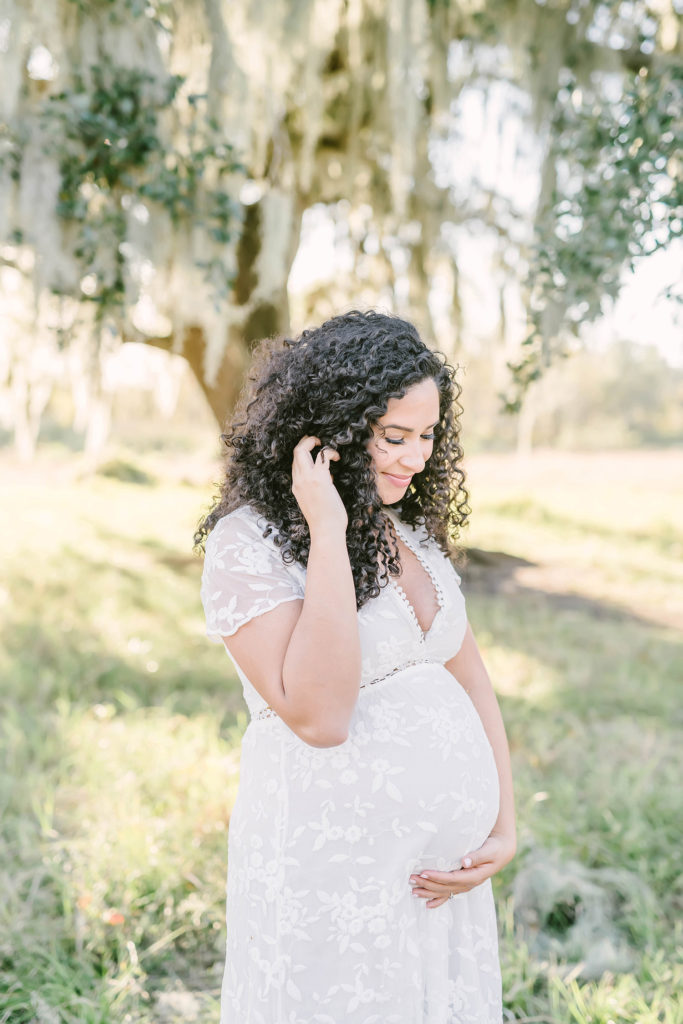 Beneath a shade tress in Brazos Bend State Park, Houston, Texas maternity photographer, Christina Elliott Photography captures this expectant mother's portrait. white v-neck lace maxi maternity dress women's full shoulder length curly hair Houston Texas winter photo session #HoustonMaternityPhotographer #HoustonCouplesPhotographer #HoustonPhotographer #MaternitySession #MaternityDresses #ChristinaElliottPhotography #HoustonFamilyPhotographer #Pregnant #BabyBump #WereExpecting