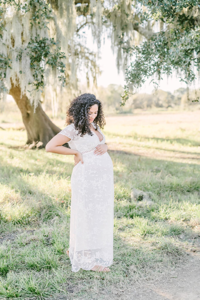 In Brazos Bend State Park, Houston, Texas maternity photographer, Christina Elliott Photography captures an up-close portrait of this expectant mother holding her baby bump. mother looking down at baby bump Houston Texas maternity photographer white lace maxi dress maternity dress #HoustonMaternityPhotographer #HoustonCouplesPhotographer #HoustonPhotographer #MaternitySession #MaternityDresses #ChristinaElliottPhotography #HoustonFamilyPhotographer #Pregnant #BabyBump #WereExpecting