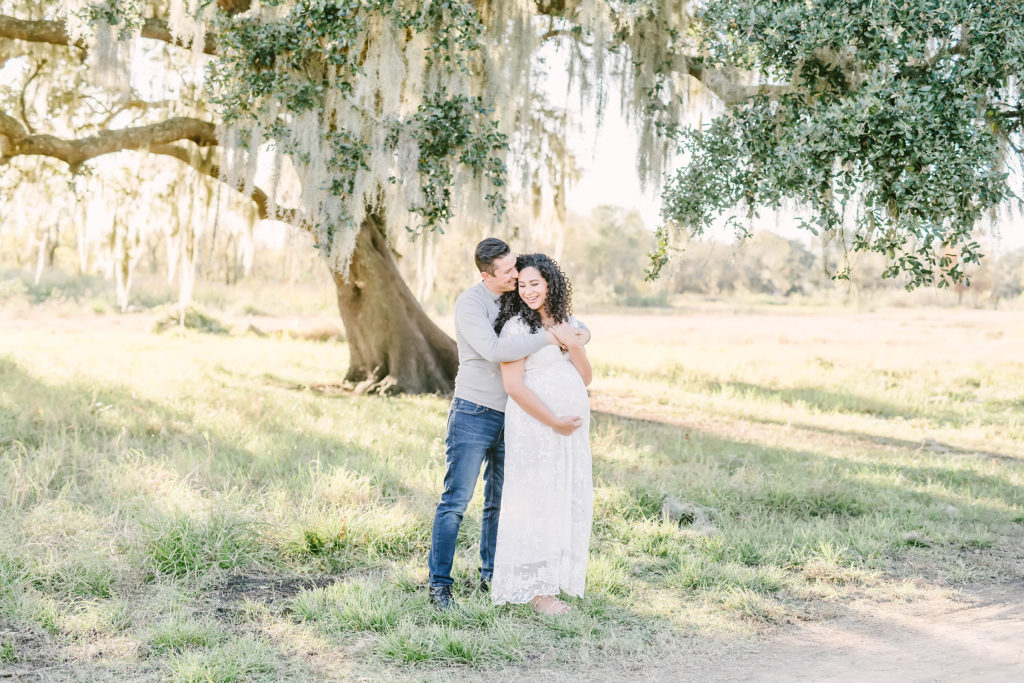 While standing under a shade tree in Houston, Texas, Christina Elliott Photography this soon-to-be father kissing his wife's cheek and cupping her baby bump. tender maternity session poses Houston Texas maternity photographer white lace cap sleeved maternity dress #HoustonMaternityPhotographer #HoustonCouplesPhotographer #HoustonPhotographer #MaternitySession #MaternityDresses #ChristinaElliottPhotography #HoustonFamilyPhotographer #Pregnant #BabyBump #WereExpecting