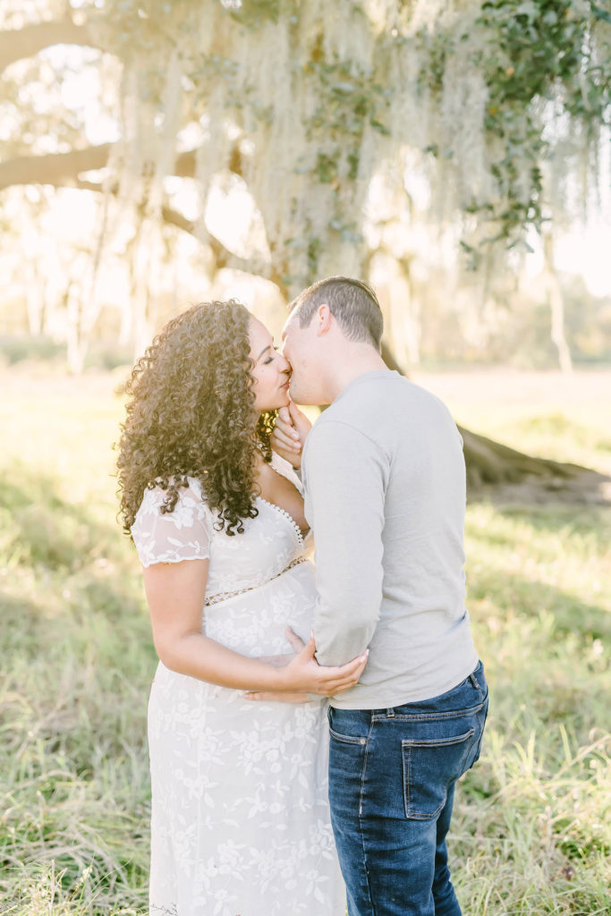 While standing under a shade tree in Houston, Texas, Christina Elliott Photography this expectant couple tenderly kissing. expectant couple kissing women's shoulder length full curly hair white lace v-neck maxi dress maternity Houston photographer session #HoustonMaternityPhotographer #HoustonCouplesPhotographer #HoustonPhotographer #MaternitySession #MaternityDresses #ChristinaElliottPhotography #HoustonFamilyPhotographer #Pregnant #BabyBump #WereExpecting