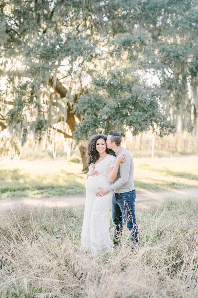 While standing under a shade tree in Houston, Texas, Christina Elliott Photography captures this expectant couple tenderly embracing and kissing. man kissing woman's cheek father hugging baby bump Houston Texas photographer women's maternity maxi dress lace #HoustonMaternityPhotographer #HoustonCouplesPhotographer #HoustonPhotographer #MaternitySession #MaternityDresses #ChristinaElliottPhotography #HoustonFamilyPhotographer #Pregnant #BabyBump #WereExpecting