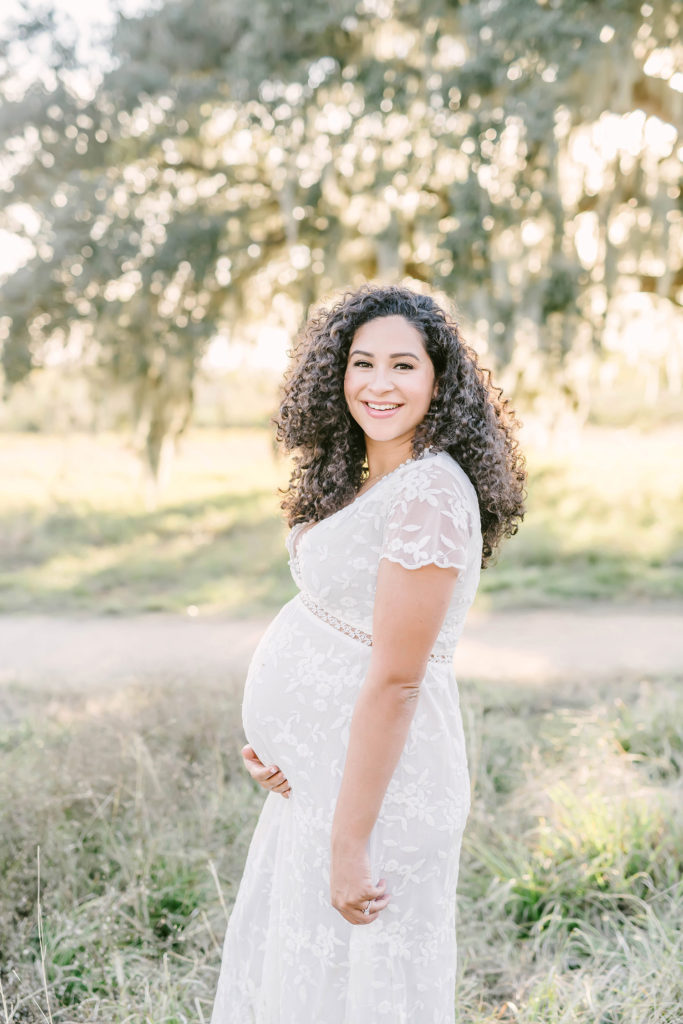 Christina Elliott Photography captures this smiling woman in a field of tall grass in Houston, Texas while she poses with her growing baby bump during her maternity session. smiling maternity session Houston photographer white short sleeve lace maxi maternity dress #HoustonMaternityPhotographer #HoustonCouplesPhotographer #HoustonPhotographer #MaternitySession #MaternityDresses #ChristinaElliottPhotography #HoustonFamilyPhotographer #Pregnant #BabyBump #WereExpecting