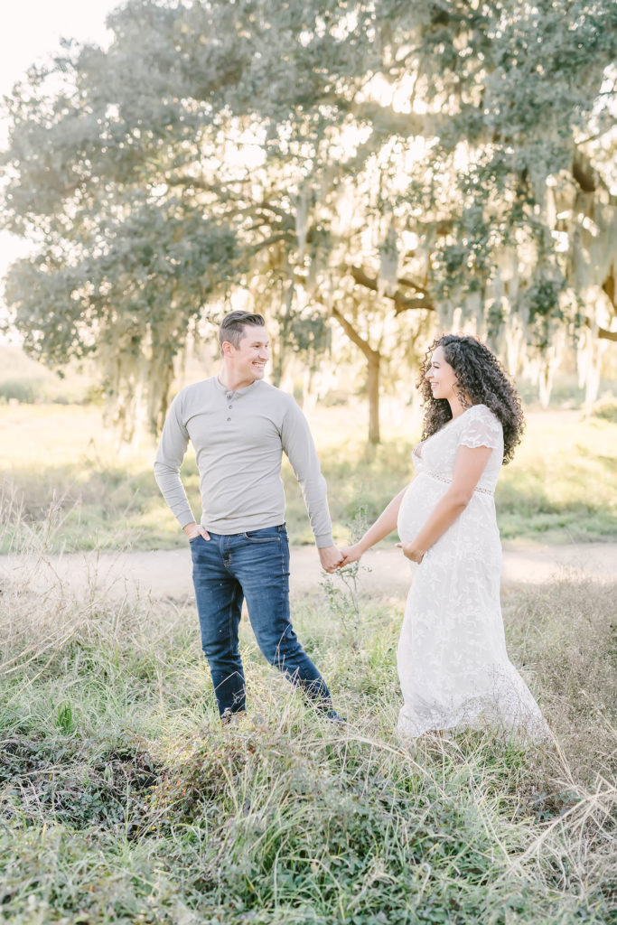 Houston, Texas maternity photographer, Christina Elliott Photography captures this expectant couple holding hands and smiling together as they walk through tall grass. tall grass playful maternity session women's lace white maxi dress baby bump holding hands couple Houston photographer #HoustonMaternityPhotographer #HoustonCouplesPhotographer #HoustonPhotographer #MaternitySession #MaternityDresses #ChristinaElliottPhotography #HoustonFamilyPhotographer #Pregnant #BabyBump #WereExpecting