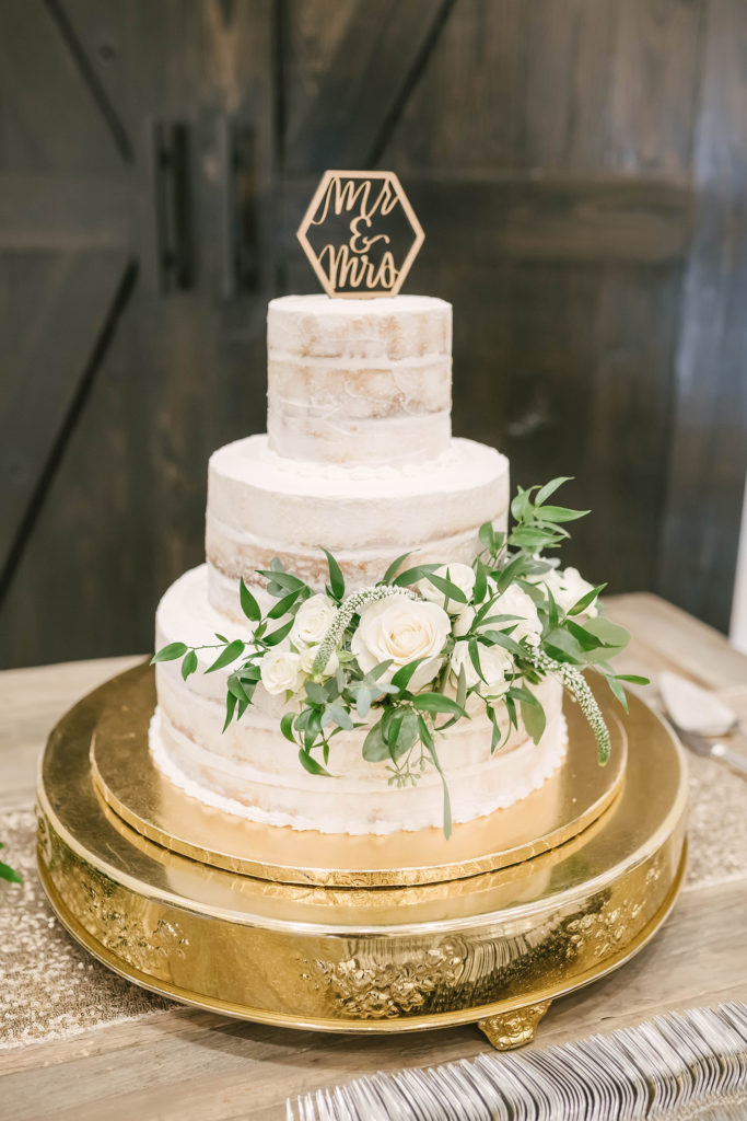 An elegant view of this simple wedding cake for a barn style wedding in Alvin, Texas captured by Christina Elliot Photography. Simple wedding cake gold cake stand greenery white cake topper Mr and Mrs barn style country wedding cake alvin texas wedding photographer #houstonweddingphotographer #texasphotographer #weddingdaydetails #barnstylewedding #stillwatersranch #alvintexasphotographer #weddingday #brideandgroom #texanwedding #htxwedding #countrywedding