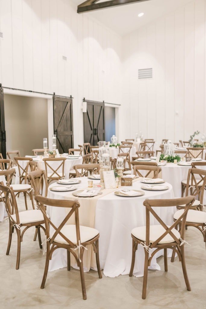 A view of the tables at a barn style wedding reception in Houston, Texas with Christina Elliot Photography. Barn style country wedding still waters ranch alvin texas wedding photographer table settings white elegant wooden chairs centerpiece inspo #houstonweddingphotographer #texasphotographer #weddingdaydetails #barnstylewedding #stillwatersranch #alvintexasphotographer #weddingday #brideandgroom #texanwedding #htxwedding #countrywedding