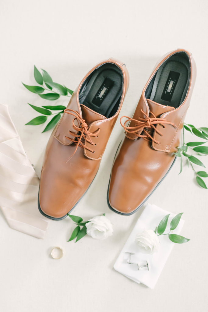 A close up shot of the grooms wear details captured by Christina Elliot Photography in Alvin, Texas. Grooms shoes brown leather white tie wedding suit inspo gold wedding ring greenery tie cufflinks details wedding day Houston texas wedding photographer #houstonweddingphotographer #texasphotographer #weddingdaydetails #barnstylewedding #stillwatersranch #alvintexasphotographer #weddingday #brideandgroom #texanwedding #htxwedding #countrywedding