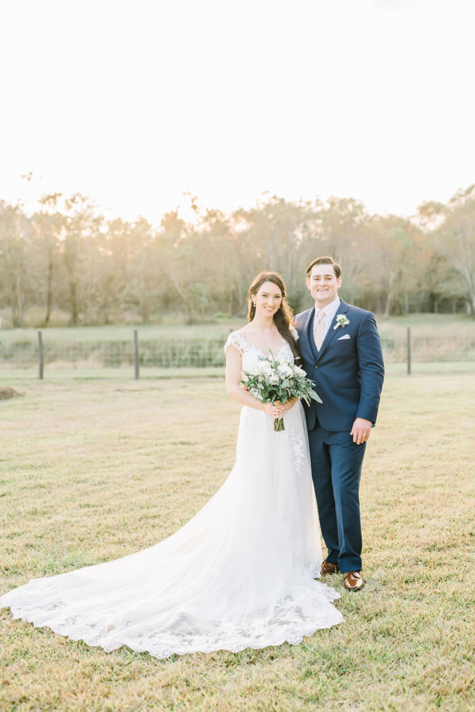This beautiful bride and groom couple pose for the camera outside their barn style wedding during bridals with Christina Elliot Photography in Houston, Texas. Smiles picture posing inspo wedding day bride and groom white lace wedding dress navy blue suit flower bridal bouquet #houstonweddingphotographer #texasphotographer #weddingdaydetails #barnstylewedding #stillwatersranch #alvintexasphotographer #weddingday #brideandgroom #texanwedding #htxwedding #countrywedding