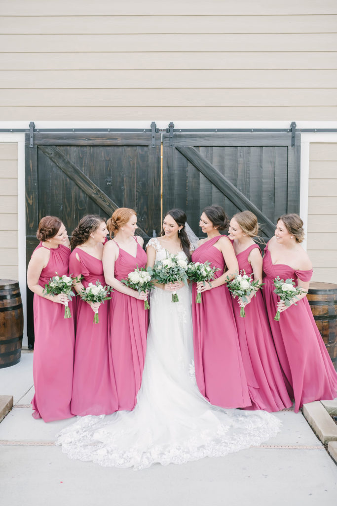 A bride and her bridesmaids in front of the barn at Still Waters Ranch in Alvin, Texas captured by Christina Elliot Photography. Bride bridesmaids white wedding dress maroon bridesmaids dress inspo flower bouquets black barn door white lace train texas wedding photographer #houstonweddingphotographer #texasphotographer #weddingdaydetails #barnstylewedding #stillwatersranch #alvintexasphotographer #weddingday #brideandgroom #texanwedding #htxwedding #countrywedding