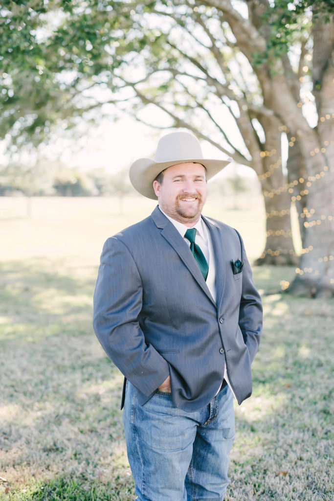 Groom smiles in grey suit with beige cowboy hat behind the barn at the Barn at Willowynn wedding venue
