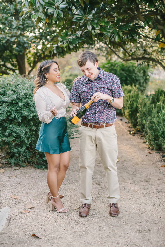 A little engagement celebration during a beautiful garden themed photoshoot in Houston, Texas with Christina Elliot Photography. Champagne flutes celebration engaged greenery backdrop outdoor springtime engagement session montgomery texas wedding photographer teal skirt white blouse printed blue button up shirt #houstonweddingphotographer #engagementpics #houstonphotographer #engagementphotos #shesaidyes #texasphotographer #photoinspiration #weddingphotography #houstonphotographer