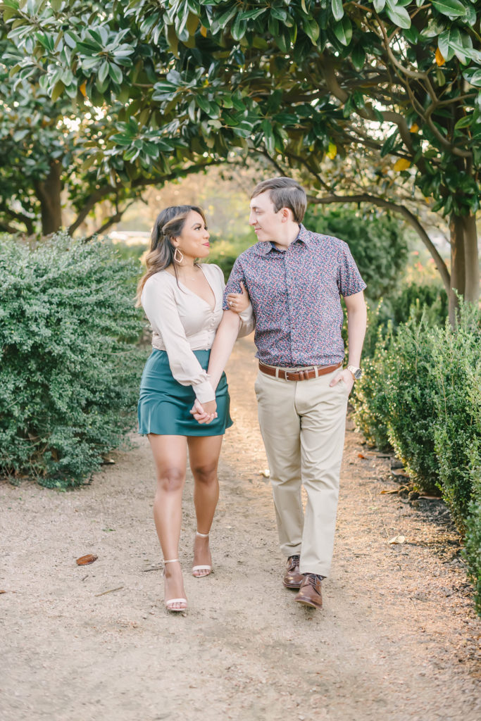 A happy couple strolls together outside during their engagement photoshoot in Houston, Texas with Christina Elliot Photography. Texas wedding photographer walking together she said yes engagement session green trees outside photoshoot semi formal attire inspo gold earrings jewelry #houstonweddingphotographer #engagementpics #houstonphotographer #engagementphotos #shesaidyes #texasphotographer #photoinspiration #weddingphotography #houstonphotographer
