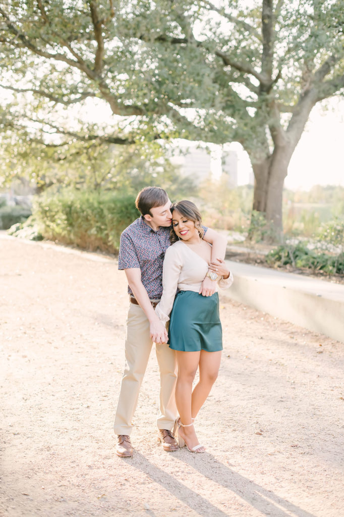 This sweet couple poses together for their engagement session with Christina Elliot Photography in Downtown Houston, Texas. Picture pose inspo montgomery texas wedding photographer teal skirt button up shirt tan pants heels smiles green trees outdoor engagement photo session #christinaelliotphotography #houstonweddingphotographer #engagementpics #houstonphotographer #engagementphotos #shesaidyes #texasphotographer #photoinspiration #weddingphotography #houstonphotographer