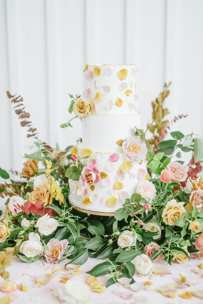 White three tier wedding cake with flower accents by Wink by Erica at the Farmhouse wedding venue surrounded by a floral design by Jackie Trejo Floral Design.