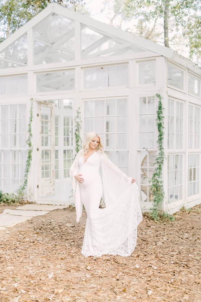 A soon to be momma at Oak Atelier holds her white country lace dress up while holding her belly bump by Christina Elliott Photography. window pane building white greenhouse #christinaelliottphotography #christinaelliotmaternitysession #babybump #houstonmaternityphotographer #houstonmaternity #pregnancypic #houstonphotographylocations #materntiyoutfit #soontobeparents #babyontheway #houstonphotos #houstontexasmaternityphotographer #oakatelierphotos #conroetexas