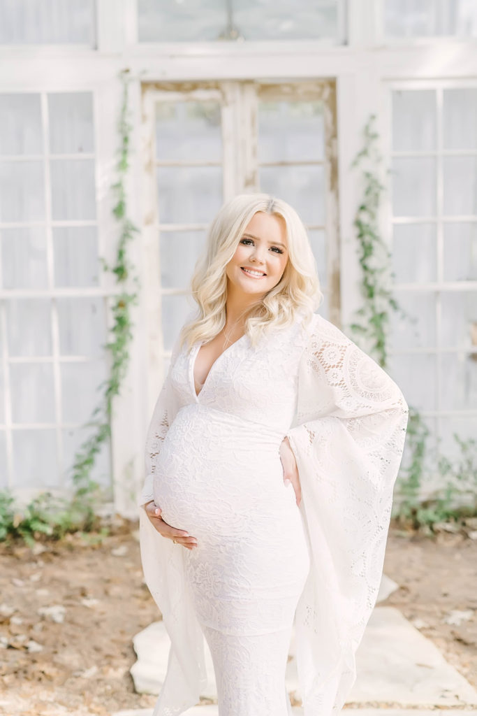 Mama smiles while holding her baby bump in white boho crochet maternity gown with bell sleeves by Christina Elliott Photography in Conroe. bell sleeve dress country style maternity pic #christinaelliottphotography #christinaelliotmaternitysession #babybump #houstonmaternityphotographer #houstonmaternity #pregnancypic #houstonphotographylocations #materntiyoutfit #soontobeparents #babyontheway #houstonphotos #houstontexasmaternityphotographer #oakatelierphotos #conroetexas