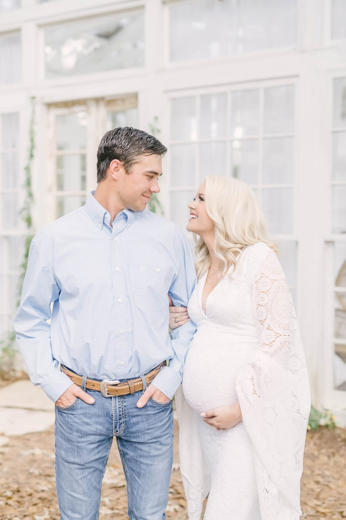 At Oak Atelier a husband and wife look at each other while woman holds her baby bump by Christina Elliott Photography. blonde woman lace sleeve dress parents to be #christinaelliottphotography #christinaelliotmaternitysession #babybump #houstonmaternityphotographer #houstonmaternity #pregnancypic #houstonphotographylocations #materntiyoutfit #soontobeparents #babyontheway #houstonphotos #houstontexasmaternityphotographer #oakatelierphotos #conroetexas