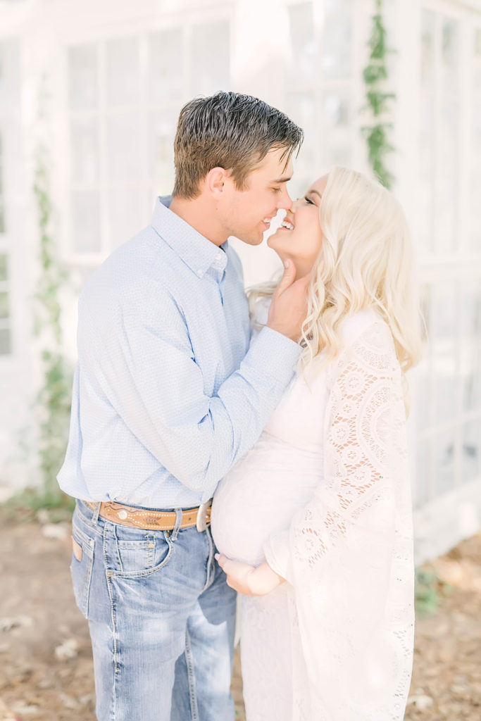 A husband and wife smile while they are kissing during a maternity session in Oak Atelier in Conroe, Texas by Christina Elliott Photography. white crochet maternity dress color scheme for maternity session #christinaelliottphotography #christinaelliotmaternitysession #babybump #houstonmaternityphotographer #houstonmaternity #pregnancypic #houstonphotographylocations #materntiyoutfit #soontobeparents #babyontheway #houstonphotos #houstontexasmaternityphotographer #oakatelierphotos #conroetexas