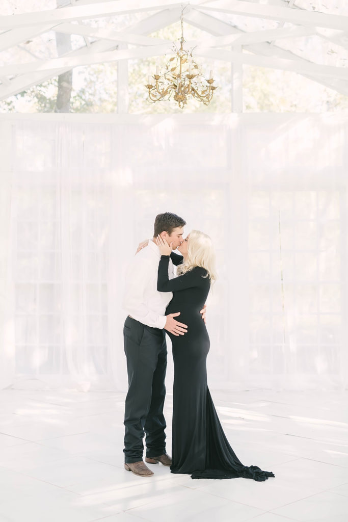 Soon-to-be mother and father kiss while man holds her belly by Christina Elliot Photography out of Houston. maternity kissing pic black maternity dress white backdrop gold chandlier #christinaelliottphotography #christinaelliotmaternitysession #babybump #houstonmaternityphotographer #houstonmaternity #pregnancypic #houstonphotographylocations #materntiyoutfit #soontobeparents #babyontheway #houstonphotos #houstontexasmaternityphotographer #oakatelierphotos #conroetexas