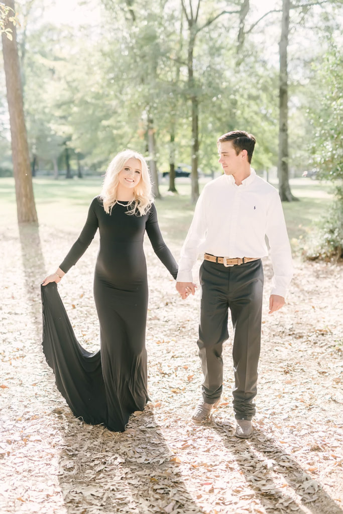A beautiful pregnant woman in a black fitted dress holds hands with her man while walking in a forest in Conroe, Texas by Christina Elliot Photography. fitted maternity dress baby bump style awaiting baby #christinaelliottphotography #christinaelliotmaternitysession #babybump #houstonmaternityphotographer #houstonmaternity #pregnancypic #houstonphotographylocations #materntiyoutfit #soontobeparents #babyontheway #houstonphotos #houstontexasmaternityphotographer #oakatelierphotos #conroetexas