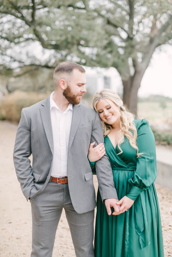 Groom lovingly looks over at his bride in her green maxi dress and holds her hand during their engagement photos with Christina Elliott Photography at Eleanor Tinsley Park in downtown Houston.
