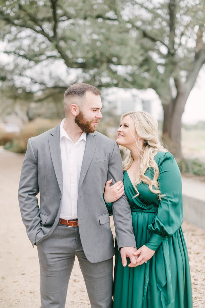 Engaged couple smile at a downtown houston park for their engagement session. Bride to be in a green maxi dress smiles up at her fiance in a gray suit.