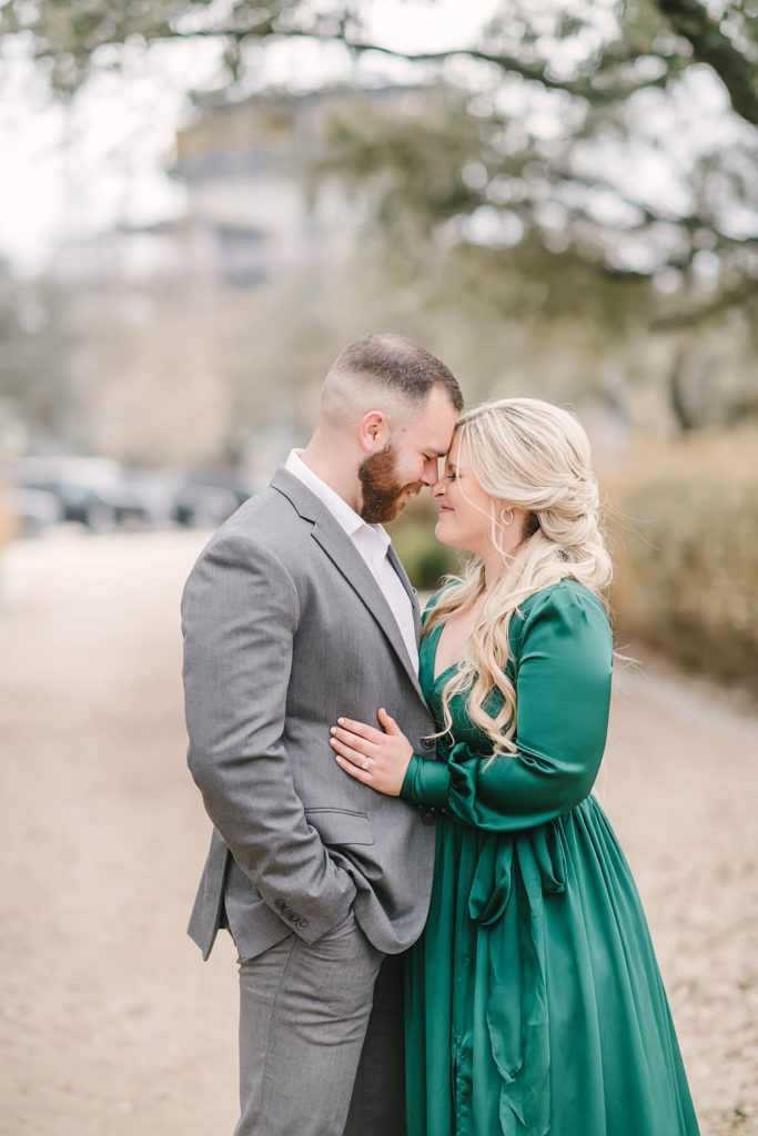 Couple cozies up face to face during photo session. Groom to be wears a grey suit to the engagement photos and bride to be wears a stunning emerald maxi dress.