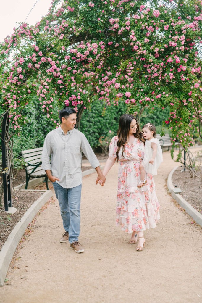 Family holds hands through the rose gardens at McGovern Centennial Gardens. Mom is wearing a flowy floral maxi dress and baby in a white dress with a pink bow.