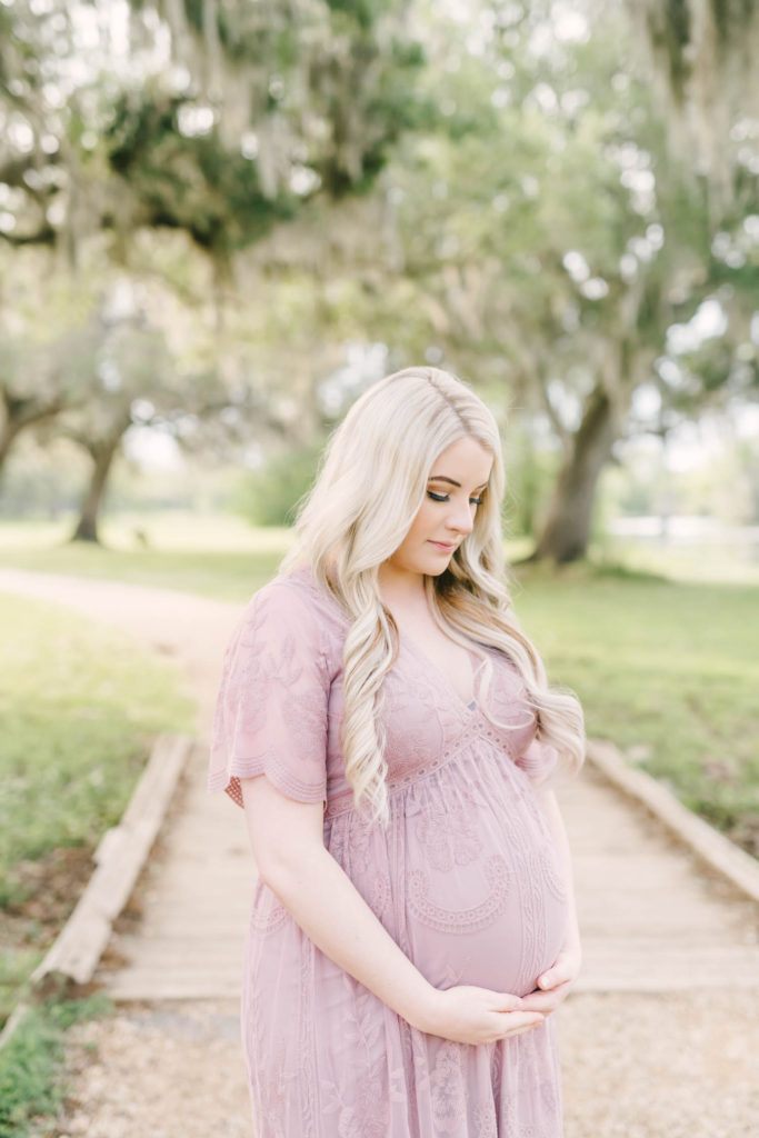 At Brazos Bend State Park in Houston a mother in a mauve floor length gown holds her pregnant belly while looking down by Christina Elliott Photography. floor length maternity dress mauve family pictures outdoor maternity #christinaeliottphotography #christinaelliotmaternity #brazosbendstatepark #houstonmaternityphotographer #parentstobe #babybumppicutes #houstonareaphotographer #professionalmaternityphotographer #houstonmaternity #preggo #pregnancyjournal #texasmaternityphotographer