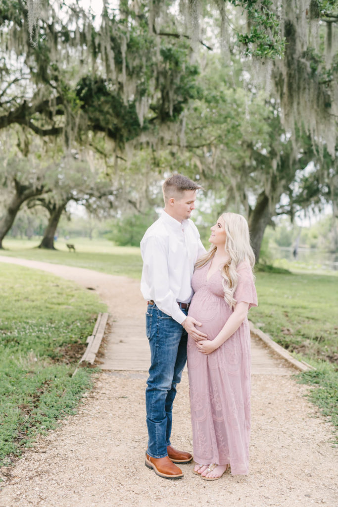 Professional Texas Maternity Photographer Christina Elliott Photography takes a picture of a nine months pregnant woman in a soft pink dress holding her belly looking at her husband in boots on a country road. country road pic soft pink dress prof photographer #christinaelliotmaternity #brazosbendstatepark #houstonmaternityphotographer #parentstobe #babybumppicutes #houstonareaphotographer #professionalmaternityphotographer #houstonmaternity #preggo #pregnancyjournal #texasmaternityphotographer