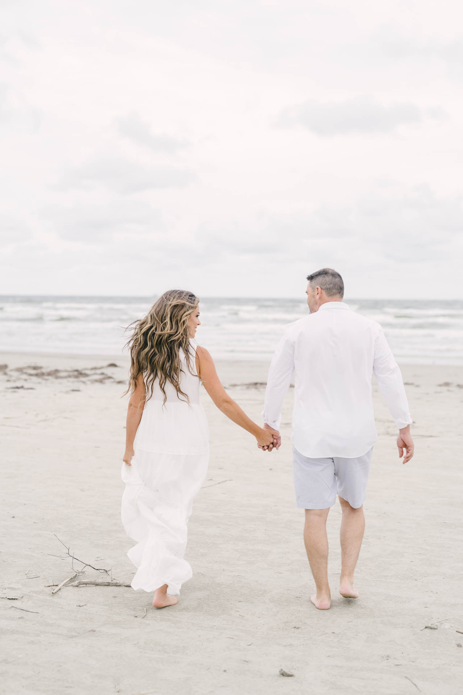 Couple walk hand in hand towards the ocean on Galveston Island for their beach engagement session with Christina Elliott Photography.