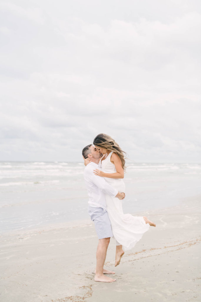 Husband lifts wife for sweet kiss during their engagement session on Galveston Island in the Beachtown community beach access.