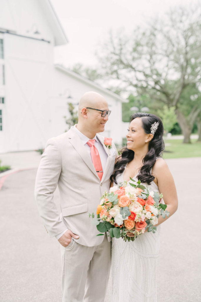 A groom wearing a white suit and coral tie looks lovingly at his bride who has beautiful black hair and a coral bouquet in Texas by Christina Elliott Photography. adoring couple elopements in Wallisville spring wedding #christinaelliotphoto #christinaelliotphotography #christinaelliotelopements #elopementphotographer #wallisvillewedding #houstontexaswedding #houstonweddingphotographer #trinityfarmhouse #texaselopements #elopement #springwedding