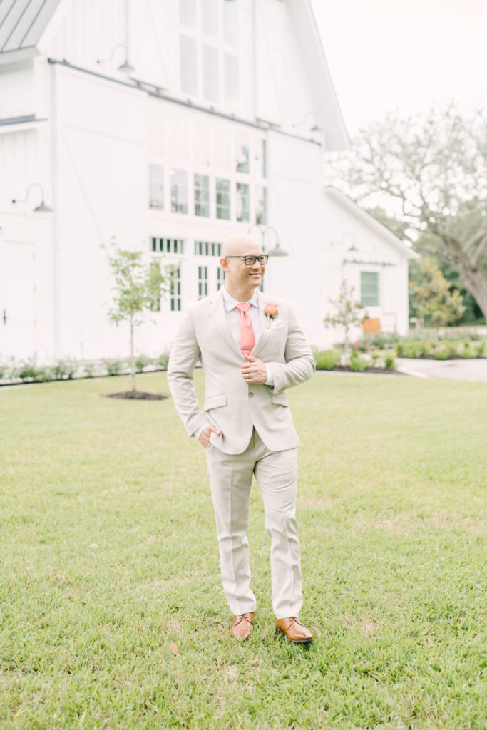 Christina Elliott Photography a Houston Wedding Photographer captures a groom portrait in front of a white barn in Texas during an elopement. groom portrait groom style inspiration brown groom shoes cream suit coral tie black glasses #christinaelliotphoto #christinaelliotphotography #christinaelliotelopements #elopementphotographer #wallisvillewedding #houstontexaswedding #houstonweddingphotographer #trinityfarmhouse #texaselopements #elopement #springwedding