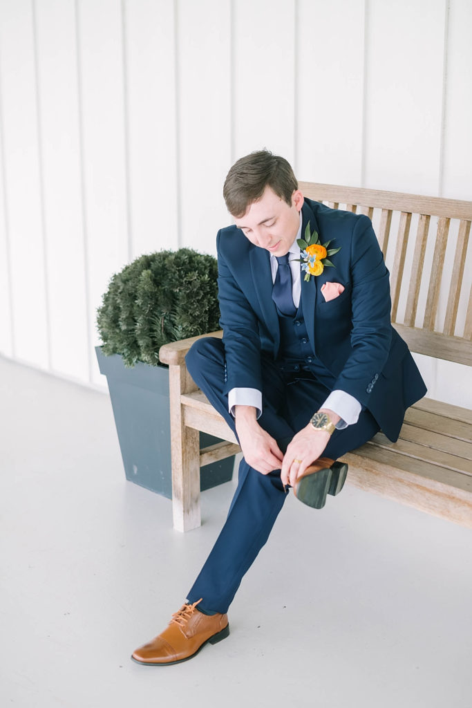 Handsome groom wearing a navy blue suit and brown shoes with a yellow flower perfect for a summer wedding by Christina Elliot Photography in Texas. how to dress your groom best suit for groom colors for groom to wear for summer wedding navy suit for groom yellow flowers for summer farmhouse wedding handsome groom for wedding #texasweddingphotographer #farmhousewedding #farmhouseweddinginspo #texasphotographer #farmhouseweddingdecor #texasweddinglocations
