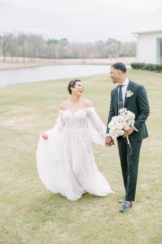 Christina Elliott Photography captures a newly wed couple walking hand-in-hand laughing at one another at The Farmhouse Montgomery. laughing bride and groom summer wedding gown floral bodice wedding dress groom style #christinaelliottphotography #christinaelliottweddings #TheFarmhouseMontgomery #farmhouseweddings #texasweddings #houstonweddingphotographers #mrandmrs #tildeathdowepart #weddingphotography #tietheknot #Houstonweddings #weddinginspiration #bride #groom